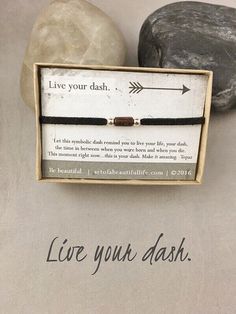 a black cord bracelet with an arrow on it in front of a rock that says live your dash