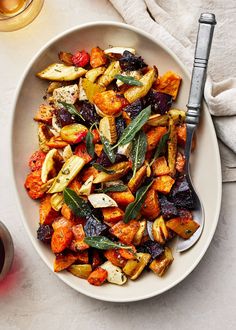 a white bowl filled with roasted vegetables on top of a table next to a glass of wine