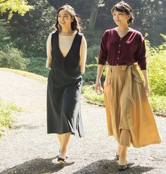 two women walking down a dirt road in the woods, one wearing a brown dress