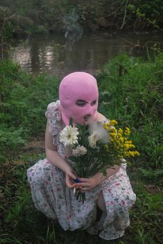 a woman wearing a pink mask holding flowers in front of her face while sitting on the ground