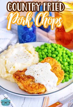 a white plate topped with fried pork chops next to green peas and mashed potatoes