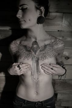 a woman with a bird tattoo on her chest is standing in front of a wooden wall