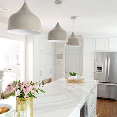 a white kitchen with marble counter tops and pendant lights over the island, along with two vases filled with flowers