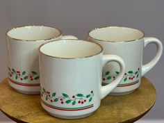 three white coffee mugs with holly designs on them sitting on a wooden table top