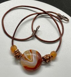 This vibrant necklace features a striking Sardonyx Agate pendant, known for its bold bands of color, complemented by warm copper and rich Carnelian beads. Strung on a durable leather cord, the natural textures and tones create a perfect balance of earthy elegance and bright, energetic design. Ideal for those who love statement pieces with a touch of artisanal flair, this necklace adds a pop of color and warmth to any outfit. Approximately 20" long. For reference Sardonyx Agate in almost 1" aroun Adjustable Wire Wrapped Carnelian Necklace, Brown Agate Hand Wrapped Necklace, Adjustable Carnelian Necklace Hand Wrapped, Unique Adjustable Carnelian Necklace, Adjustable Hand Wrapped Carnelian Necklace, Artisan Brown Necklace With Waxed Cord, Amber Adjustable Hand Wrapped Necklace, Adjustable Carnelian Round Pendant Necklace, Adjustable Brown Wire Wrapped Necklace