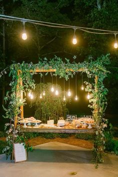 an outdoor wedding reception setup with greenery and hanging lightbulbs at night