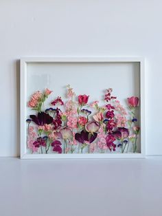 a white frame with pink and purple flowers in it on a table next to a wall