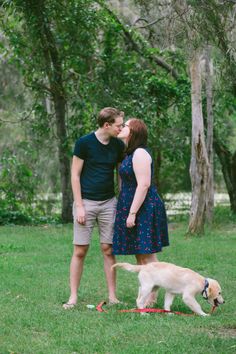 a man and woman kissing while standing next to a dog on a leash in the grass