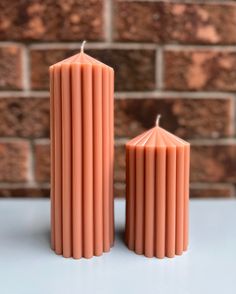 two orange candles sitting next to each other on a white table with brick wall in the background