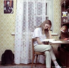 a woman sitting at a desk in front of a bookcase and window with curtains