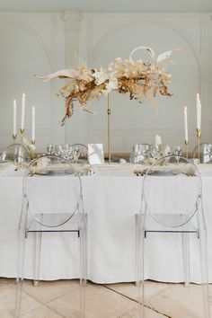 two clear chairs sitting at a table with flowers and candles in front of them on top of a white table cloth