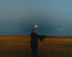 a woman standing in the middle of a field under a full moon with her arms spread out