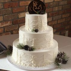 a three tiered white cake with pine cones and evergreens on the top, sitting on a table in front of a brick wall