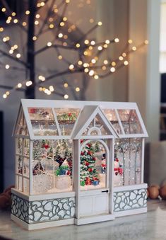 a white christmas house on a table with lights in the background and a tree behind it