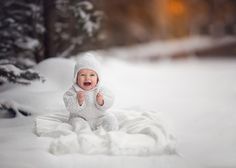 a baby is sitting in the snow wearing a white outfit and holding his thumb up