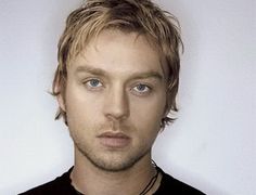 a man with blonde hair and blue eyes looks at the camera while wearing a black shirt