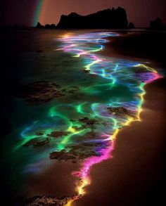 a rainbow - colored stream of water on the beach at night with a rainbow in the background