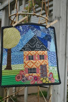 a quilt hanging from a wooden frame with a house on it's side and trees in the background