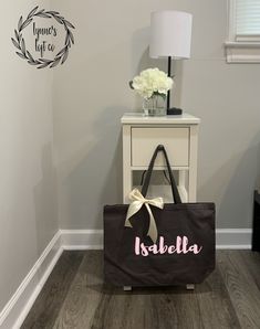 a personalized shopping bag sitting on the floor next to a nightstand with flowers in it