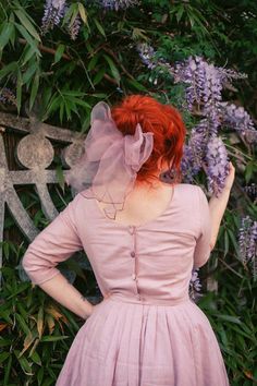 a woman with red hair wearing a pink dress and veil standing in front of purple flowers