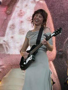 a woman in grey dress holding a guitar and smiling at the camera while standing on stage