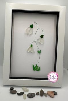 a white frame with some green and white flowers in the middle, surrounded by rocks