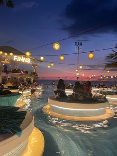 people are sitting in the middle of an outdoor swimming pool at night with lights hanging above