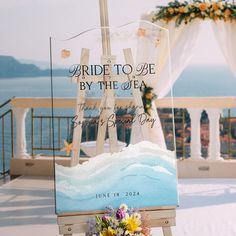 an easel with flowers on it in front of the ocean and wedding ceremony sign