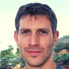 a close up of a person wearing a gray shirt and smiling at the camera with trees in the background