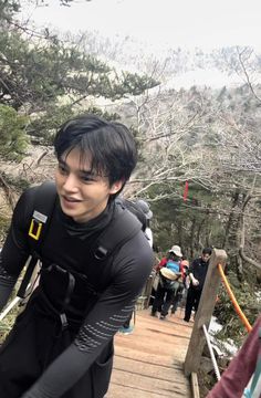 a man in black wetsuit walking down a wooden walkway next to trees and people