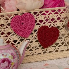 three crocheted hearts are placed next to a pink tea pot and flower arrangement