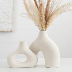 two white vases sitting next to each other with dried grass in the top one