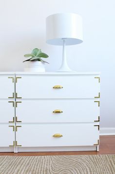 a white dresser with gold handles and a lamp on top, in front of a wall