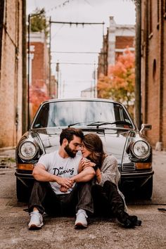 a man and woman sitting on the ground next to a car in an alleyway