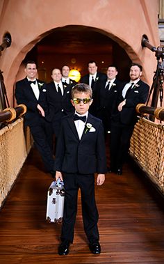 a young boy in a tuxedo is holding a suitcase and standing on a bridge