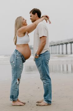 a pregnant couple standing on the beach with their arms around each other