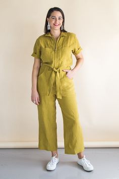 a woman standing in front of a white wall wearing a yellow jumpsuit and white sneakers