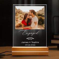 an acrylic photo frame with the words engaged on it and a couple kissing