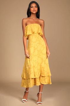 a woman wearing a yellow dress with ruffled hems and white shoes, standing in front of a beige background