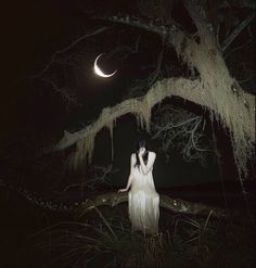 a woman in white dress standing under a tree at night with the moon behind her