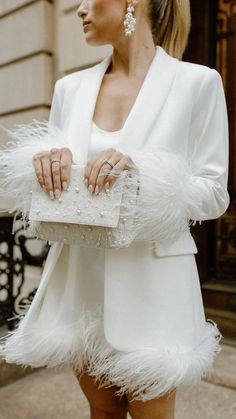 a woman in a white dress and jacket holding a white purse with feathers on it