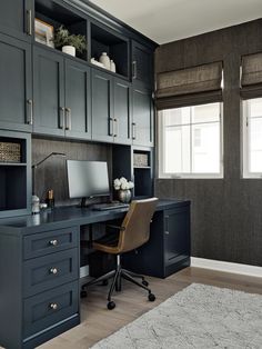 a desk with a computer on top of it in front of two windows and a rug