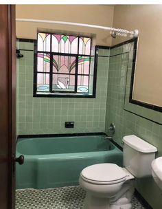 a bathroom with green tiles and a white toilet next to a bathtub in front of a stained glass window