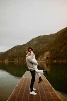 a man and woman hugging on a dock by the water with mountains in the background