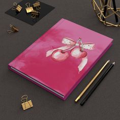 a pink notebook with scissors on it next to some gold and black items, including pens