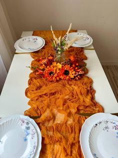 the table is set with plates and flowers on it, along with an orange runner