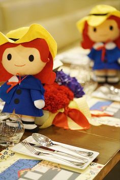 two little dolls sitting on top of a table next to silverware and napkins