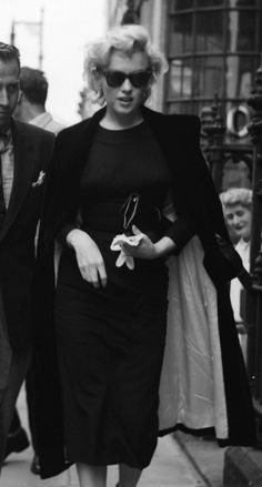 a black and white photo of a woman walking down the street with her hand in her pocket