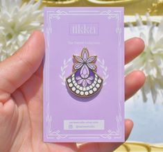 a hand holding a purple and white pin with a flower on it's back