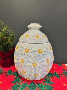 a white vase sitting on top of a table next to a potted pine tree